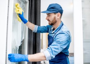 Man as a professional cleaner in blue uniform washing window with cotton wiper indoors